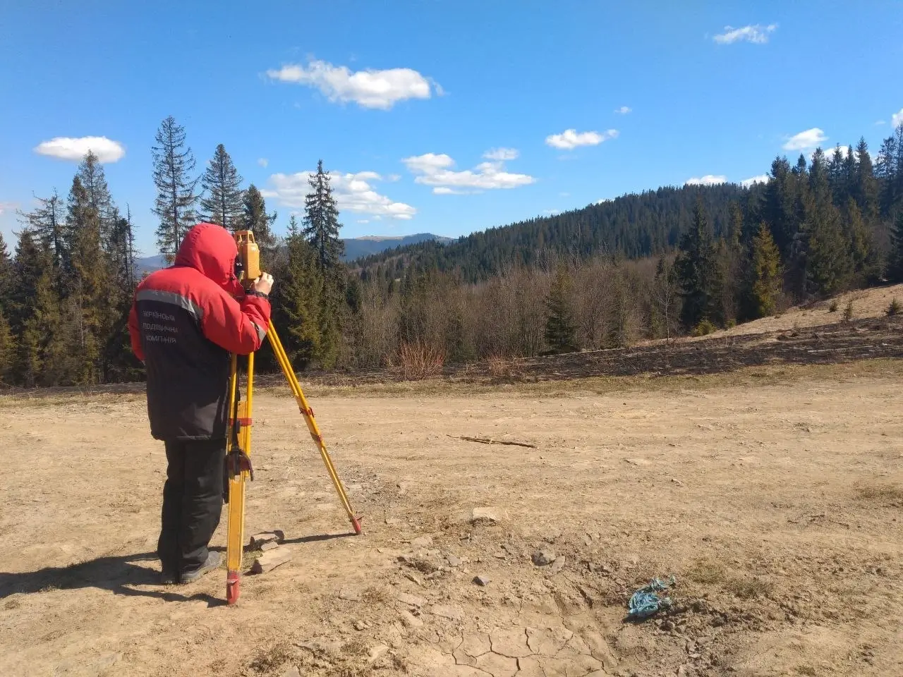 Геодезист у червоній куртці проводить виконавчу зйомку з теодолітом на відкритій місцевості серед лісу та пагорбів під ясним небом - професійні гоедезичні послуги
